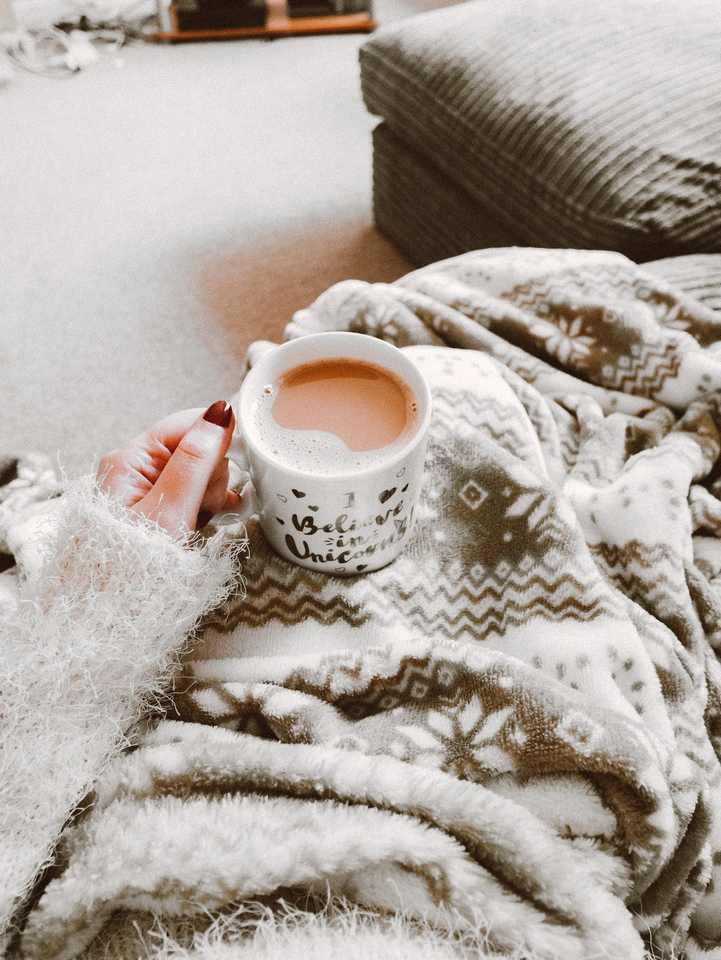 Woman's hand with red nail polish holding a cup of coffee above a blanket covering her lap.