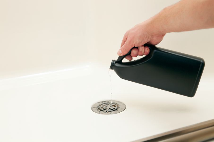 Hand pouring liquid drain cleaner from a black bottle down a shower drain.